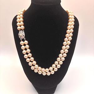 May include: A double-strand necklace featuring cream-colored round pearls of uniform size. The necklace is secured with a silver-toned metal clasp that is circular in shape and decorated with small, clear, sparkling stones set in a floral pattern.