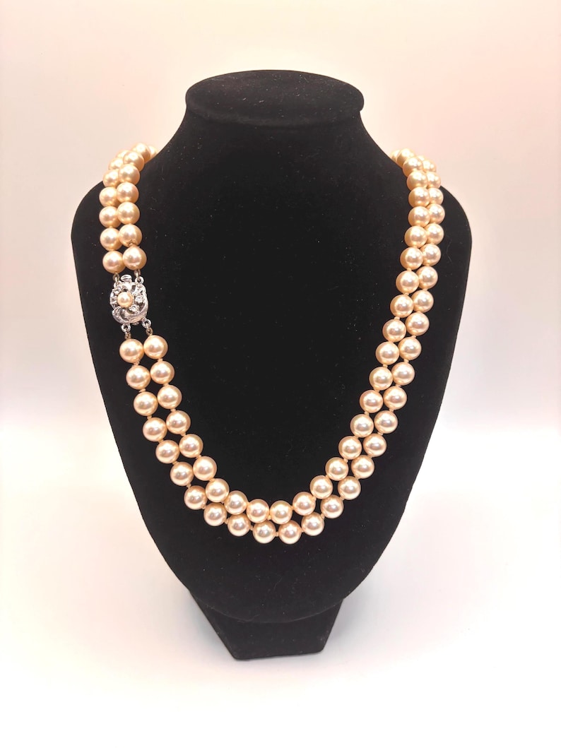 May include: A double-strand necklace featuring cream-colored round pearls of uniform size. The necklace is secured with a silver-toned metal clasp that is circular in shape and decorated with small, clear, sparkling stones set in a floral pattern.