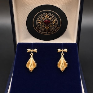 May include: A pair of gold drop earrings featuring a bow-shaped top and a textured, petal-like teardrop base. The earrings are displayed inside a dark blue velvet-lined presentation box with a circular logo reading Heirloom Splendor.
