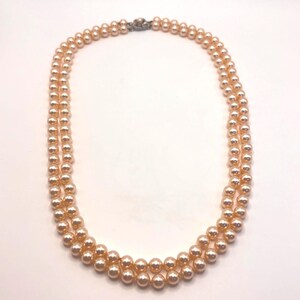 May include: A double-strand necklace featuring uniform, round, peach-colored pearls with a lustrous finish. The two strands are connected at the top by a small, metallic, decorative clasp that secures the necklace in a circular arrangement.