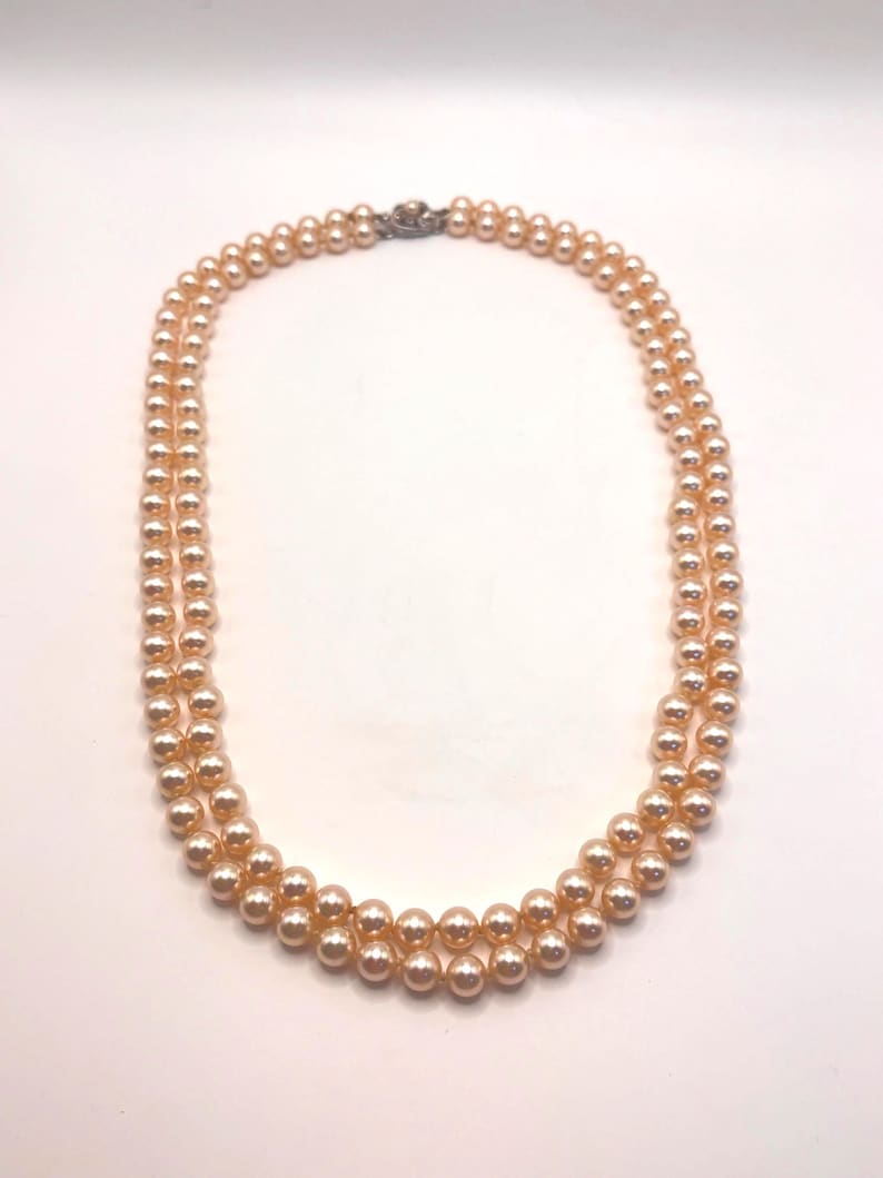 May include: A double-strand necklace featuring uniform, round, peach-colored pearls with a lustrous finish. The two strands are connected at the top by a small, metallic, decorative clasp that secures the necklace in a circular arrangement.