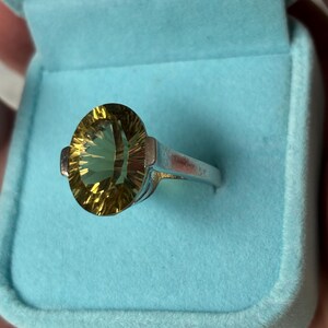 Pu&ograve; includere: Un anello color argento con una grande pietra preziosa gialla dal taglio ovale e superficie sfaccettata. L'anello &egrave; esposto all'interno di un portagioie foderato in velluto azzurro, che mette in risalto il contrasto tra la fascia metallica e la pietra.