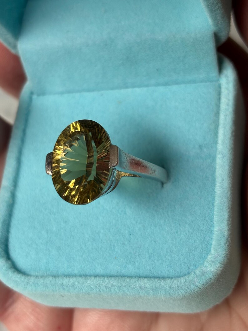 Pu&ograve; includere: Un anello color argento con una grande pietra preziosa gialla dal taglio ovale e superficie sfaccettata. L'anello &egrave; esposto all'interno di un portagioie foderato in velluto azzurro, che mette in risalto il contrasto tra la fascia metallica e la pietra.