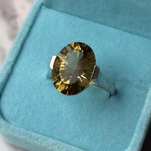 Pu&ograve; includere: Un anello in argento con una grande pietra preziosa giallo-verde dal taglio ovale e sfaccettato. La pietra presenta un motivo radiale di riflessi luminosi sulla superficie. Il gambo dell'anello &egrave; liscio e metallico, inserito in un cofanetto blu.