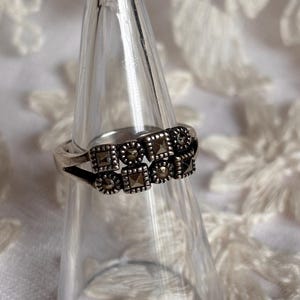 Pu&ograve; includere: Un anello d'argento con un design a doppia fascia. L'anello presenta pietre di marcasite quadrate e rotonde disposte in un motivo geometrico. L'anello &egrave; esposto su un supporto trasparente a forma di cono su sfondo bianco.