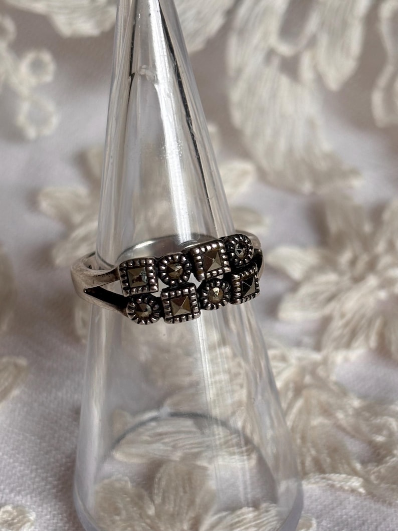 Pu&ograve; includere: Un anello d'argento con un design a doppia fascia. L'anello presenta pietre di marcasite quadrate e rotonde disposte in un motivo geometrico. L'anello &egrave; esposto su un supporto trasparente a forma di cono su sfondo bianco.