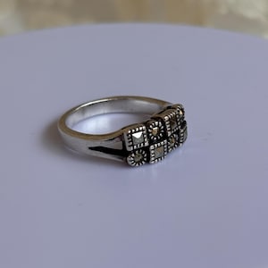 Pu&ograve; includere: Anello argentato con design rettangolare e piccole pietre quadrate. L'anello presenta uno stile vintage, con un motivo dettagliato sulla fascia. Presentato su una superficie bianca.
