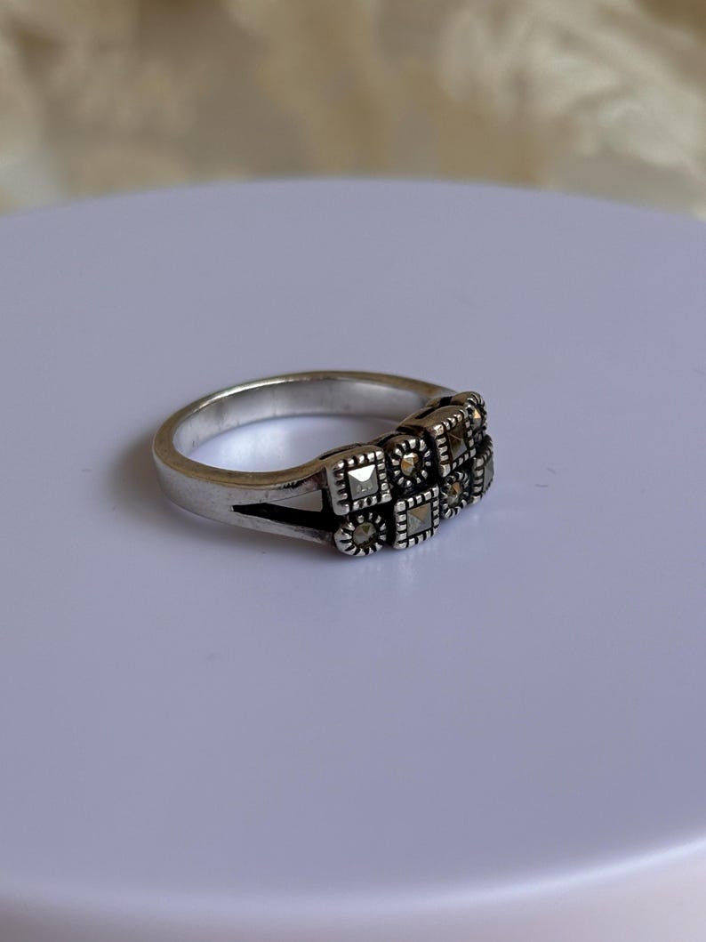 Pu&ograve; includere: Anello argentato con design rettangolare e piccole pietre quadrate. L'anello presenta uno stile vintage, con un motivo dettagliato sulla fascia. Presentato su una superficie bianca.