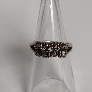 Pu&ograve; includere: Un anello d'argento con un design a doppia fascia. L'anello presenta due file di pietre di marcasite quadrate e rotonde. L'anello &egrave; esposto su un supporto trasparente a forma di cono su sfondo bianco.