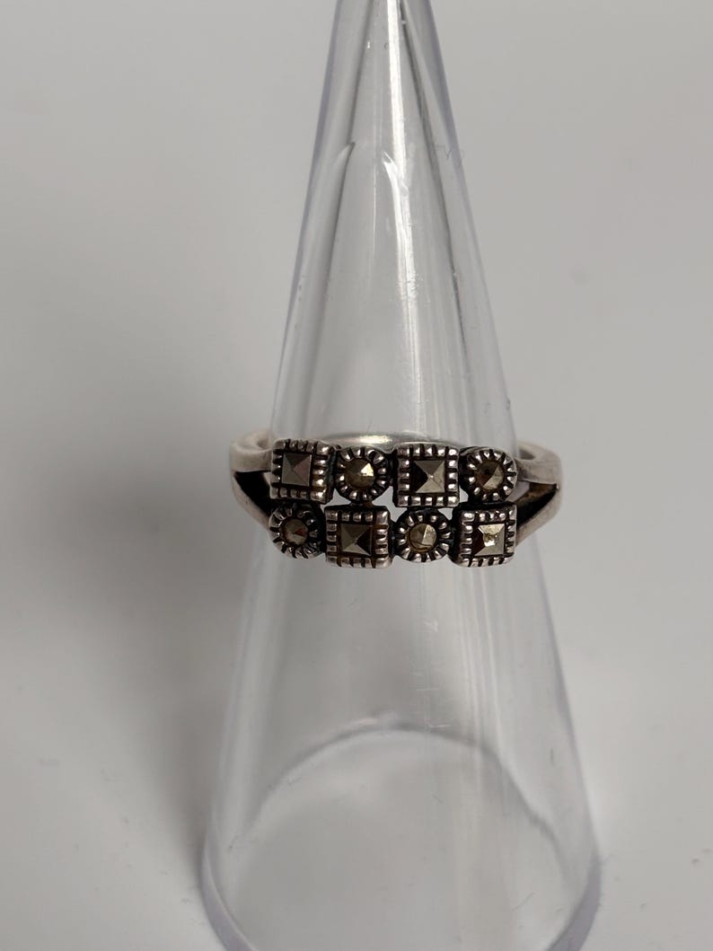 Pu&ograve; includere: Un anello d'argento con un design a doppia fascia. L'anello presenta due file di pietre di marcasite quadrate e rotonde. L'anello &egrave; esposto su un supporto trasparente a forma di cono su sfondo bianco.