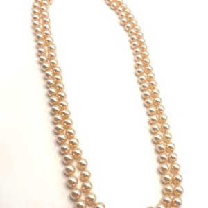 May include: A double-strand necklace featuring uniform, round, peach-colored pearls. The two strands are connected by a silver-toned metal clasp that is circular in shape and decorated with small, clear, sparkling stones arranged in a floral pattern.