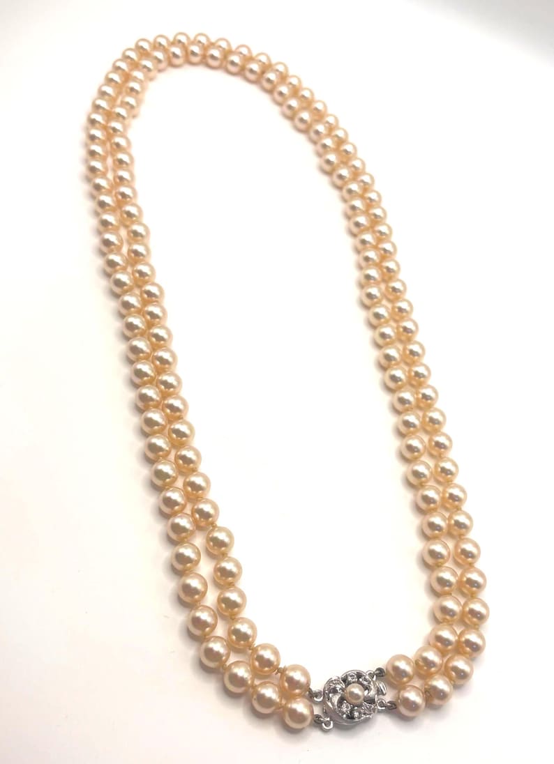 May include: A double-strand necklace featuring uniform, round, peach-colored pearls. The two strands are connected by a silver-toned metal clasp that is circular in shape and decorated with small, clear, sparkling stones arranged in a floral pattern.