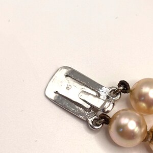 May include: A silver-toned metal box clasp for a necklace, featuring a rectangular shape with a small engraved 925 hallmark. The clasp is attached to two strands of lustrous, round, cream-colored pearls strung on dark brown thread.