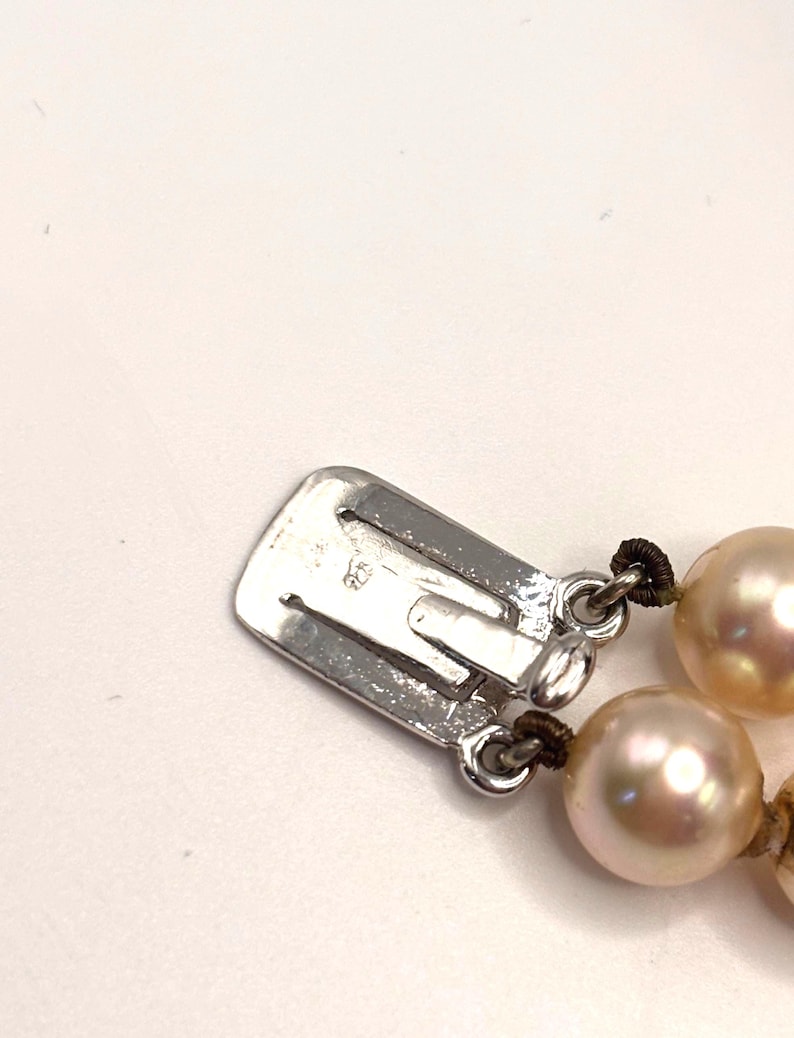May include: A silver-toned metal box clasp for a necklace, featuring a rectangular shape with a small engraved 925 hallmark. The clasp is attached to two strands of lustrous, round, cream-colored pearls strung on dark brown thread.