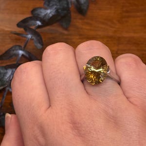 Pu&ograve; includere: Un anello in argento con una grande pietra preziosa giallo oro, taglio ovale e superficie sfaccettata. La pietra &egrave; incastonata in una semplice fascia metallica che la sostiene con griffe visibili sui lati, offrendo un aspetto chiaro e riflettente.