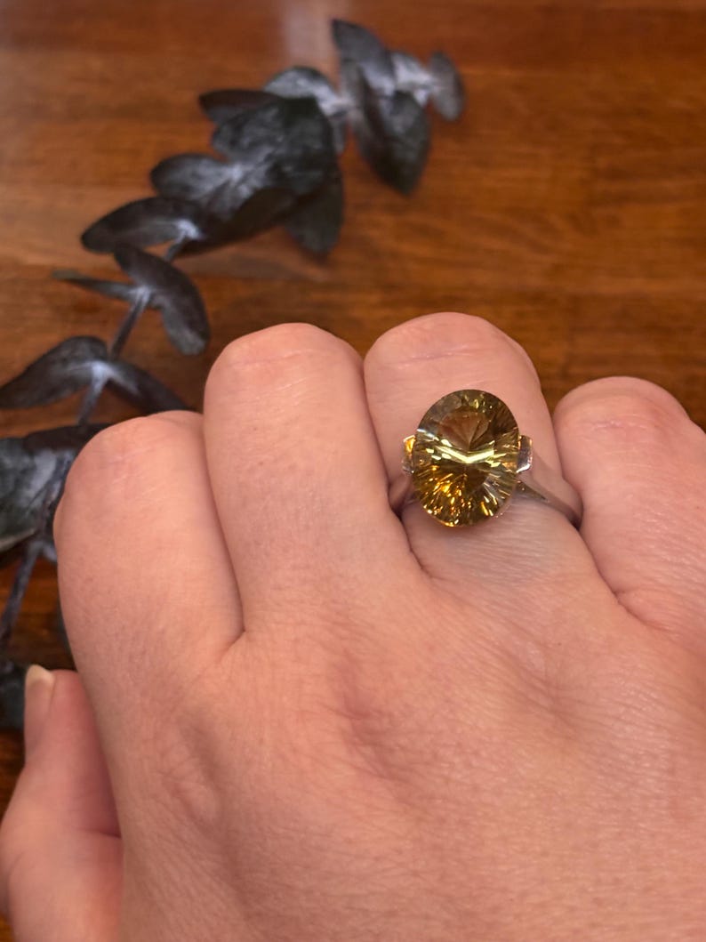 Pu&ograve; includere: Un anello in argento con una grande pietra preziosa giallo oro, taglio ovale e superficie sfaccettata. La pietra &egrave; incastonata in una semplice fascia metallica che la sostiene con griffe visibili sui lati, offrendo un aspetto chiaro e riflettente.