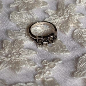 Pu&ograve; includere: Un anello d'argento con un design rettangolare con piccole pietre quadrate. L'anello &egrave; posto su un tessuto bianco a motivi floreali. La fascia dell'anello &egrave; liscia e lucida, in contrasto con le pietre strutturate.