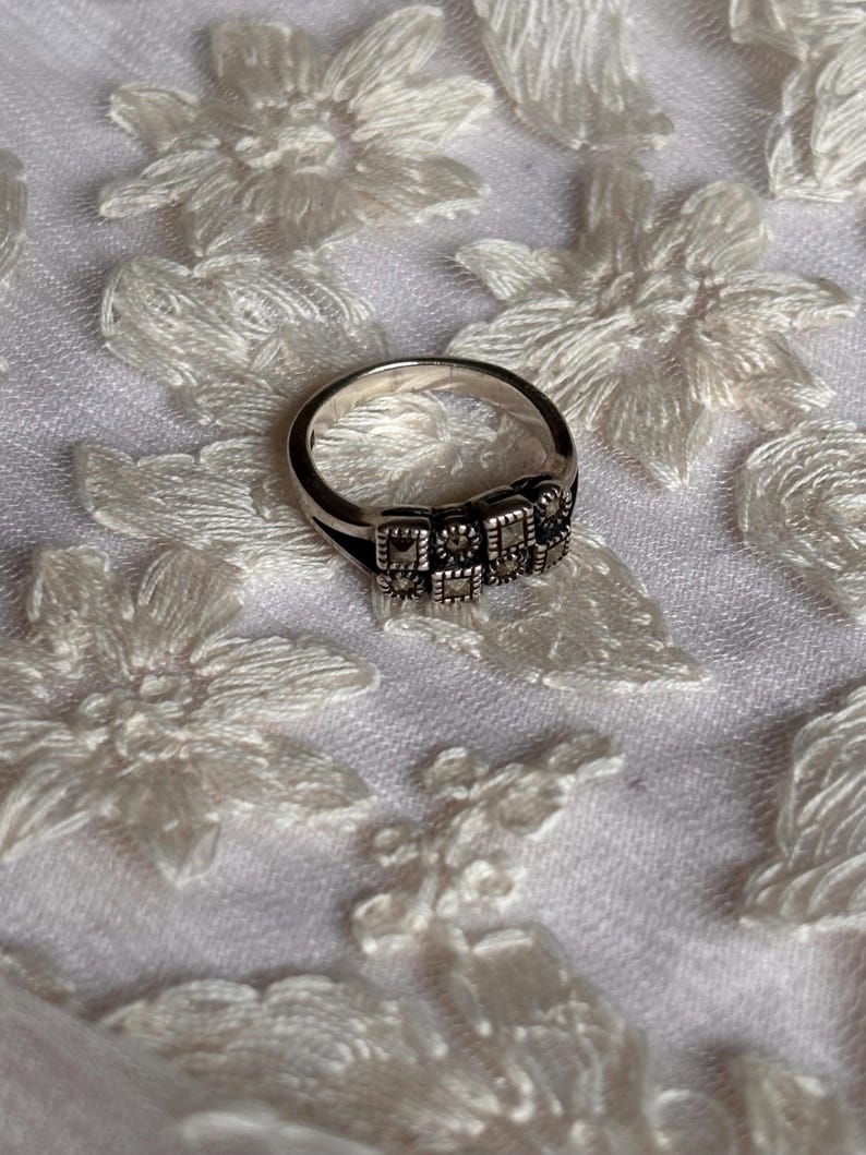 Pu&ograve; includere: Un anello d'argento con un design rettangolare con piccole pietre quadrate. L'anello &egrave; posto su un tessuto bianco a motivi floreali. La fascia dell'anello &egrave; liscia e lucida, in contrasto con le pietre strutturate.