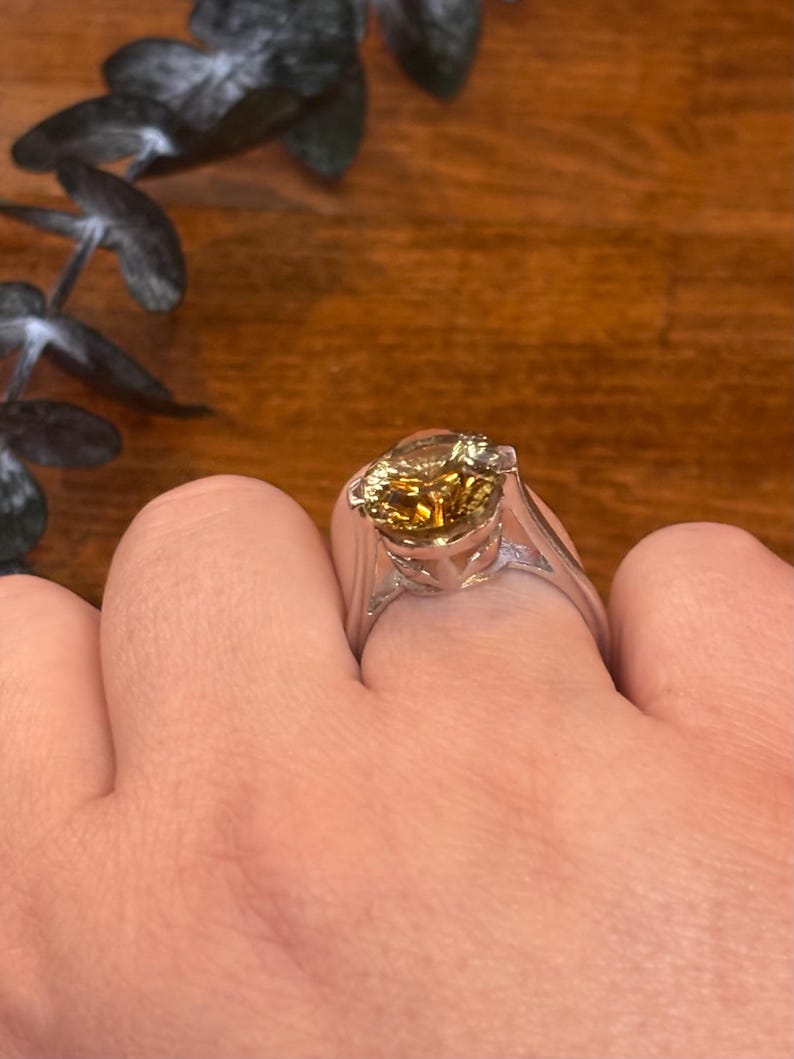 Pu&ograve; includere: Un anello color argento con una grande pietra preziosa rotonda e sfaccettata color ambra montata su una griffa. La fascia &egrave; liscia e solida, caratterizzata da una finitura metallica lucida che riflette la luce sulla pietra centrale.
