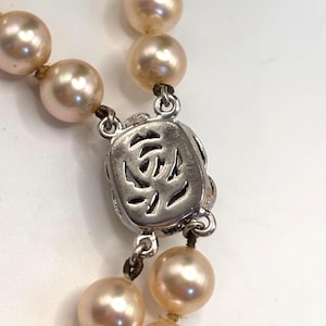 May include: A necklace featuring round, light peach-colored pearls strung on a dark cord. The necklace includes a rectangular, silver-toned metal clasp with an intricate, openwork cutout design that resembles a stylized floral or abstract pattern.