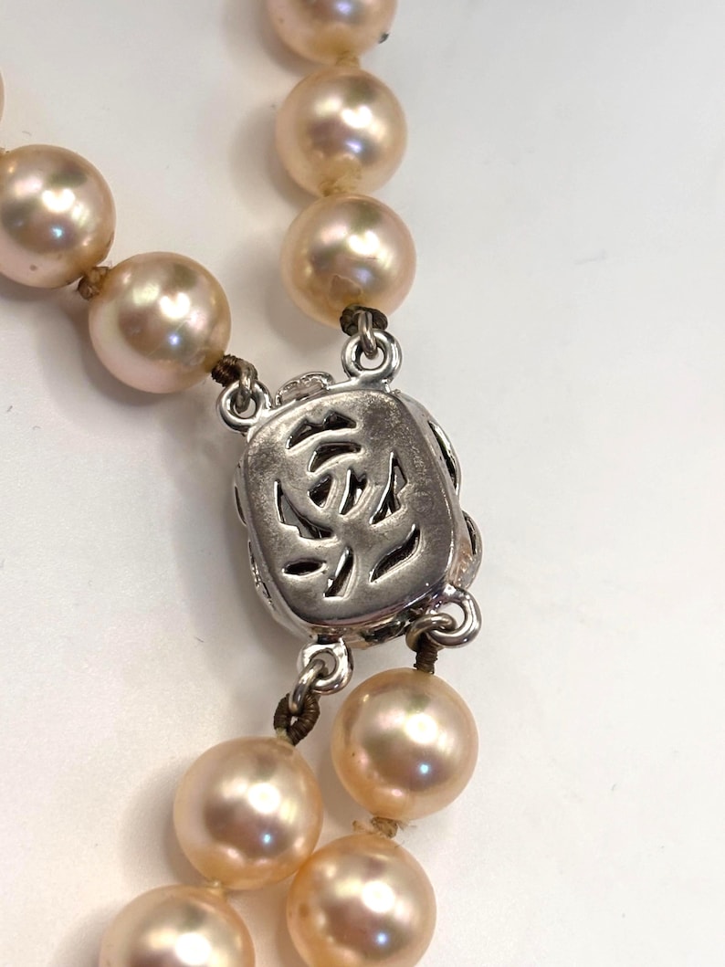 May include: A necklace featuring round, light peach-colored pearls strung on a dark cord. The necklace includes a rectangular, silver-toned metal clasp with an intricate, openwork cutout design that resembles a stylized floral or abstract pattern.