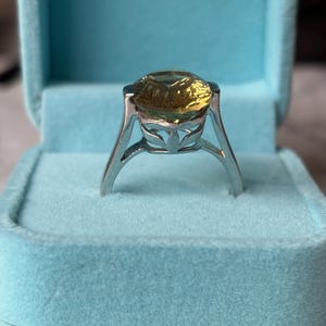Pu&ograve; includere: Un anello in argento con una grande pietra preziosa gialla rotonda e sfaccettata, incastonata in una montatura alta con dettagli traforati a forma di foglia lungo la galleria. L'anello &egrave; esposto in verticale in un portagioie azzurro foderato in velluto.