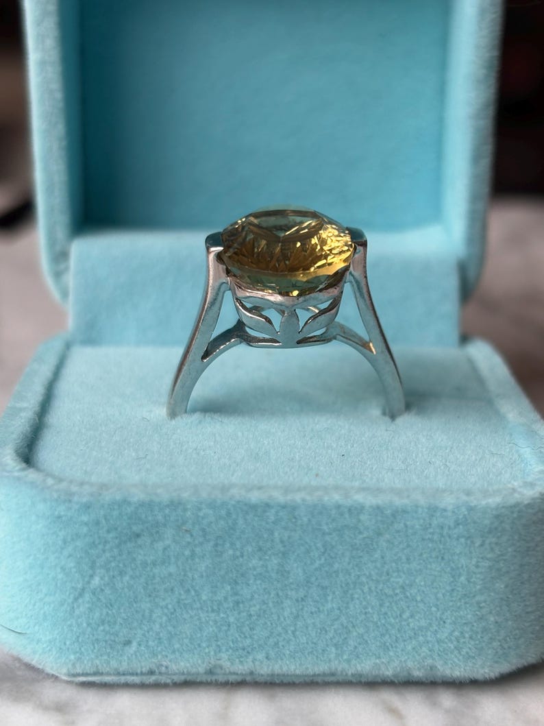 Pu&ograve; includere: Un anello in argento con una grande pietra preziosa gialla rotonda e sfaccettata, incastonata in una montatura alta con dettagli traforati a forma di foglia lungo la galleria. L'anello &egrave; esposto in verticale in un portagioie azzurro foderato in velluto.