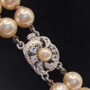 May include: A double-strand necklace featuring cream-colored pearls strung on thread. The necklace includes a silver-toned metal clasp with an intricate filigree design, set with small clear stones and a single central pearl accent.
