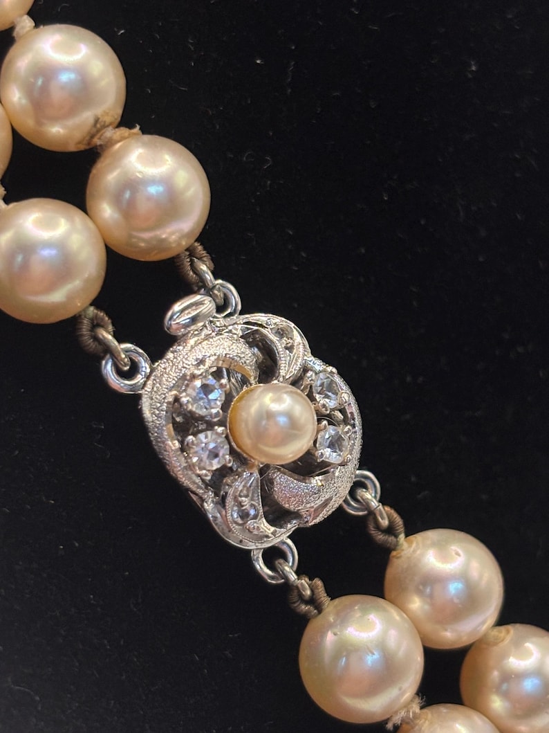 May include: A double-strand necklace featuring cream-colored pearls strung on thread. The necklace includes a silver-toned metal clasp with an intricate filigree design, set with small clear stones and a single central pearl accent.
