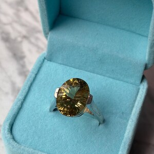 Pu&ograve; includere: Un anello in argento con una grande pietra preziosa giallo oro, taglio ovale e sfaccettature prominenti. L'anello &egrave; presentato all'interno di un portagioie azzurro foderato in velluto. La pietra preziosa mostra un motivo radiante e solare.