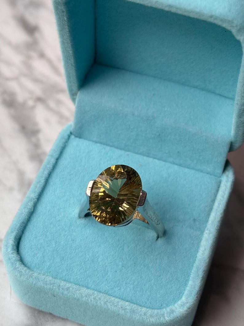 Pu&ograve; includere: Un anello in argento con una grande pietra preziosa giallo oro, taglio ovale e sfaccettature prominenti. L'anello &egrave; presentato all'interno di un portagioie azzurro foderato in velluto. La pietra preziosa mostra un motivo radiante e solare.