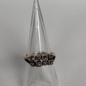 Pu&ograve; includere: Anello d'argento con una fila di elementi decorativi quadrati e rotondi. L'anello &egrave; esposto su un supporto trasparente a forma di cono, su sfondo bianco. Gli elementi decorativi hanno un aspetto scuro e strutturato.