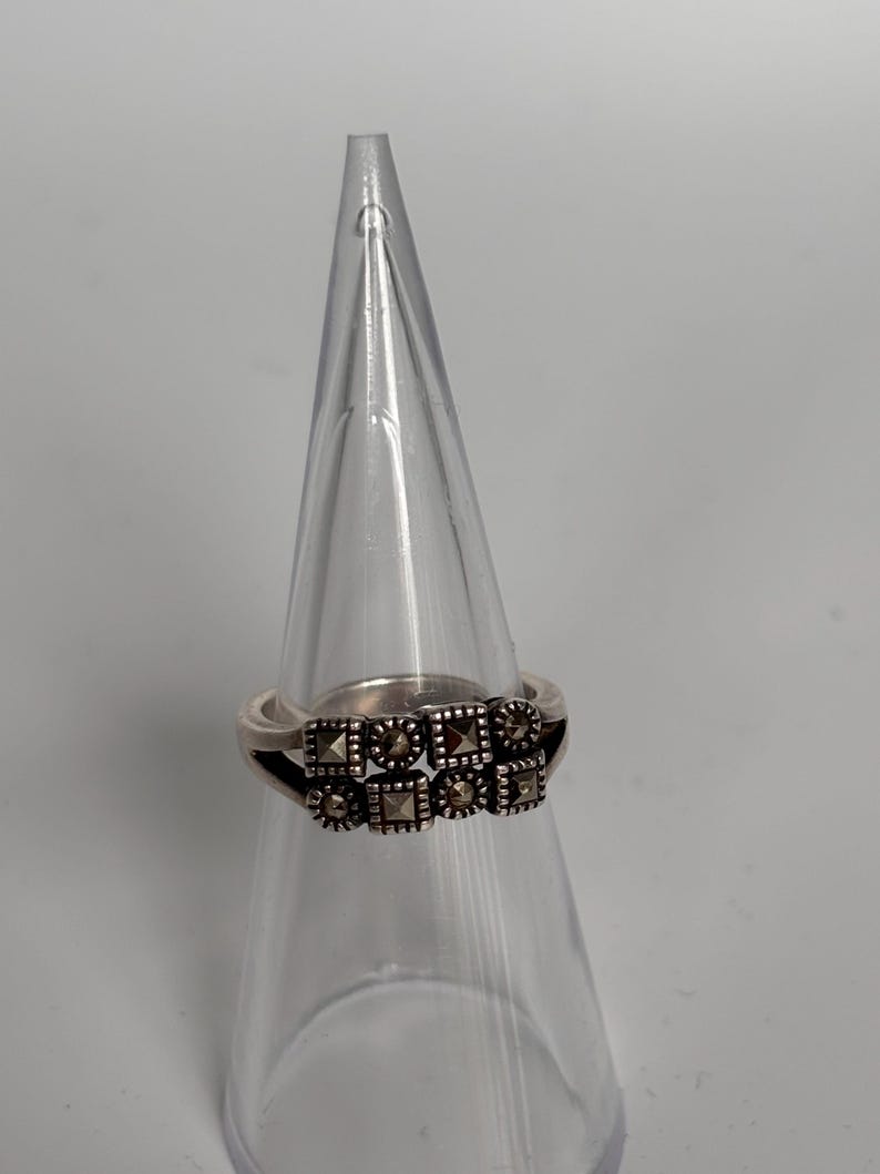 Pu&ograve; includere: Anello d'argento con una fila di elementi decorativi quadrati e rotondi. L'anello &egrave; esposto su un supporto trasparente a forma di cono, su sfondo bianco. Gli elementi decorativi hanno un aspetto scuro e strutturato.
