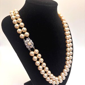 May include: A double-strand necklace featuring cream-colored pearls of uniform size. The necklace is secured with a decorative silver-toned clasp that is circular in shape and set with small, clear, sparkling stones and a single central pearl.