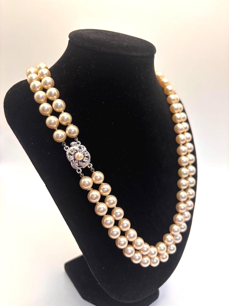 May include: A double-strand necklace featuring cream-colored pearls of uniform size. The necklace is secured with a decorative silver-toned clasp that is circular in shape and set with small, clear, sparkling stones and a single central pearl.