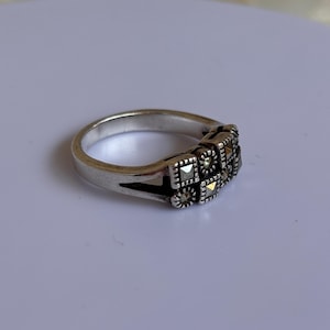 Pu&ograve; includere: Un anello d'argento con un design rettangolare con piccole pietre tagliate quadrate. L'anello ha una fascia semplice e una sezione decorativa con pi&ugrave; pietre. Le pietre sono disposte in un motivo quadrato.