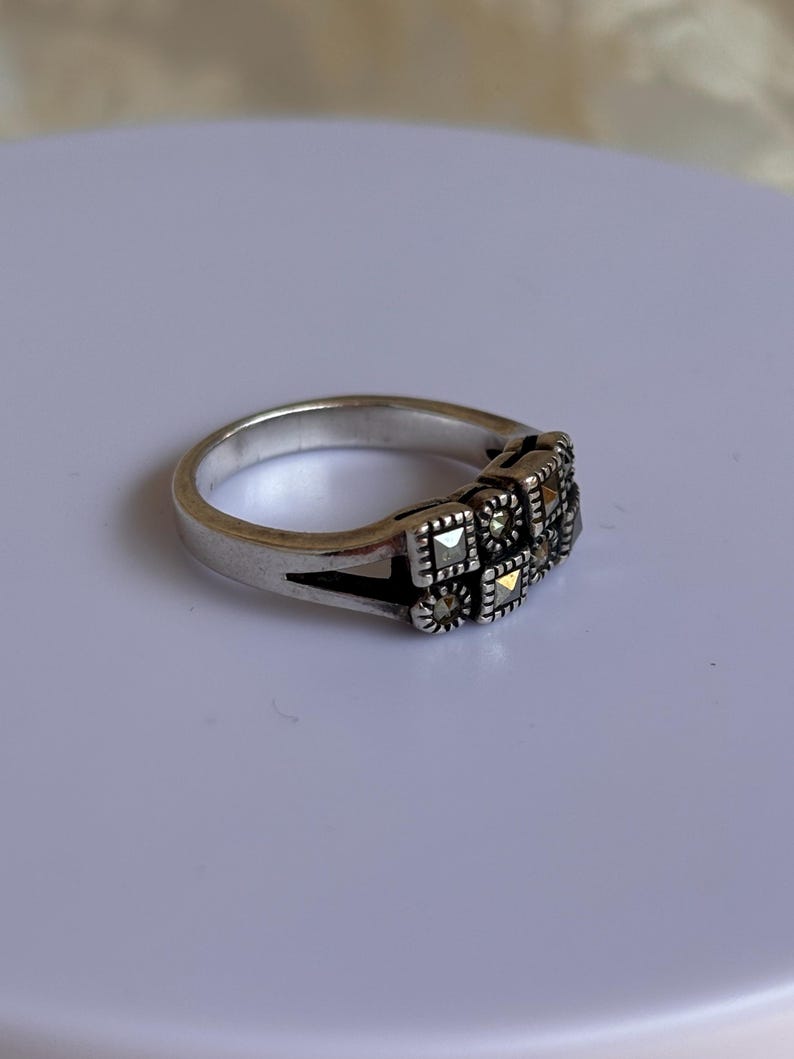 Pu&ograve; includere: Un anello d'argento con un design rettangolare con piccole pietre tagliate quadrate. L'anello ha una fascia semplice e una sezione decorativa con pi&ugrave; pietre. Le pietre sono disposte in un motivo quadrato.