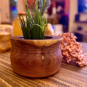 May include: A wooden planter with a dark brown finish, holding artificial plants with green grass-like leaves and purple flower heads. The planter sits on a wooden table, with a blurred background.
