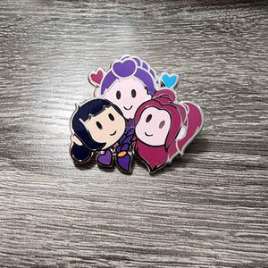 May include: A colorful enamel pin featuring three cartoon characters with smiling faces. The characters have dark blue, purple, and pink hair, with heart-shaped accents. The pin is set against a wooden background.