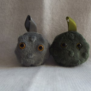 May include: Two plush toys shaped like round, furry creatures with large, round, orange eyes. The toy on the left is gray with a gray ribbon on top. The toy on the right is dark green with a green ribbon on top.