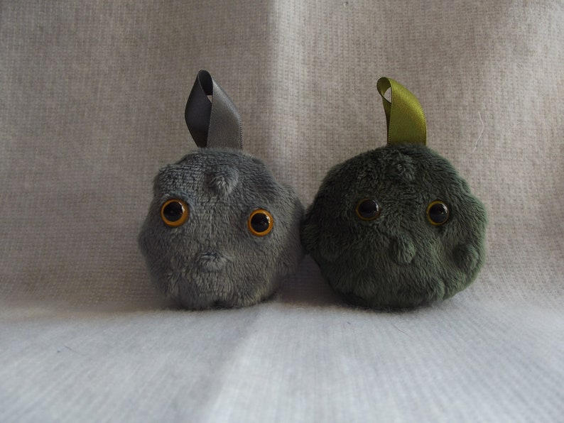 May include: Two plush toys shaped like round, furry creatures with large, round, orange eyes. The toy on the left is gray with a gray ribbon on top. The toy on the right is dark green with a green ribbon on top.
