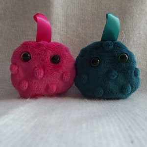 May include: Two plush toys shaped like cells, one pink and one teal blue, with black eyes and a ribbon on top.