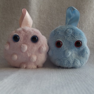 May include: Two plush toys, one pink and one blue, with large black eyes and a fuzzy texture. The pink toy has a pink ribbon on top.