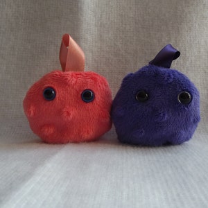 May include: Two plush toys shaped like cells. One is pink with blue eyes and the other is purple with black eyes.