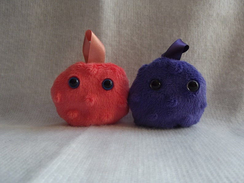 May include: Two plush toys shaped like cells. One is pink with blue eyes and the other is purple with black eyes.
