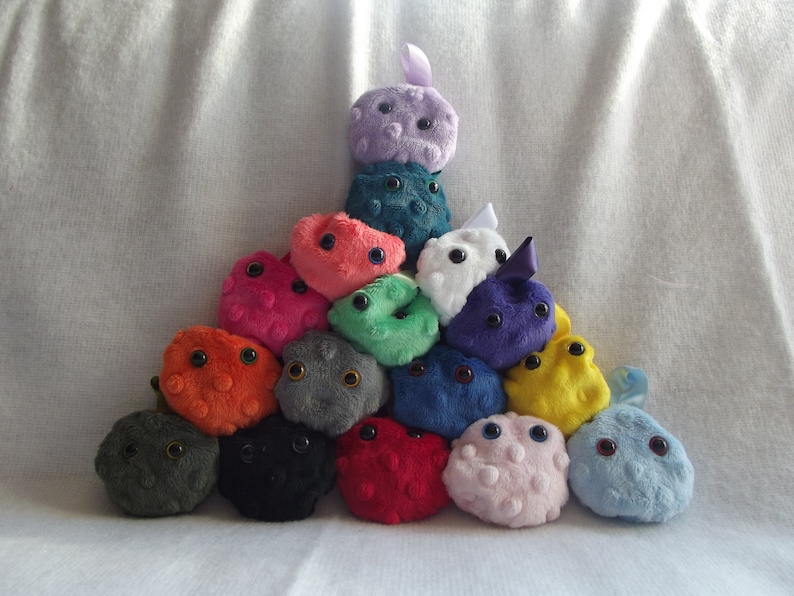 May include: A collection of 15 plush toys shaped like cells, arranged in a pyramid shape. The toys are made of soft, colorful fabric and have embroidered eyes. The colors include purple, teal, red, pink, green, yellow, orange, gray, black, and blue.