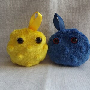 May include: Two plush toys shaped like cells. One is yellow with black eyes and the other is blue with red eyes.
