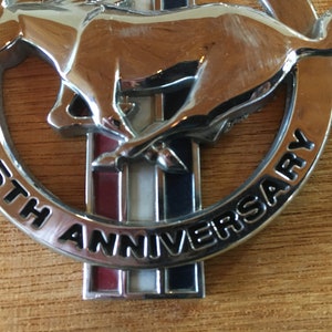 May include: A chrome-finished emblem featuring a galloping horse, the iconic Mustang logo. The emblem is circular, with the words "35TH ANNIVERSARY" in black lettering. The background has red, white, and blue stripes.