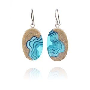 May include: A pair of oval-shaped earrings with a beach scene design. The earrings feature a light brown sand-like texture with a blue wave pattern.