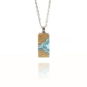 May include: A silver chain necklace with a rectangular pendant featuring a light blue and white wave design embedded in tan sand.